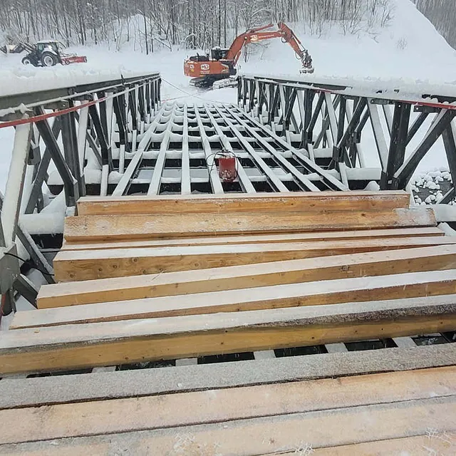 Snødekt bro under arbeid med maskiner i bakgrunnen og elv som renner under konstruksjon.