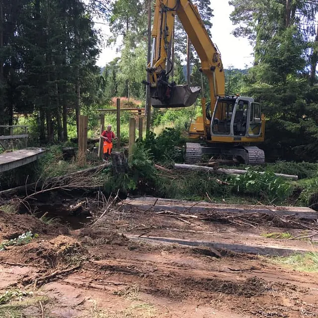 Gravemaskin arbeider ved bro og bekk i skogsterreng, med ryddearbeid og tømmer rundt.