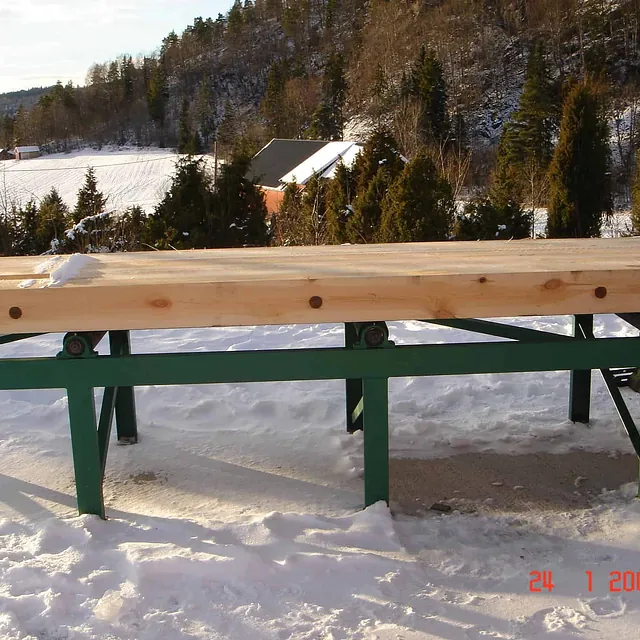 Lang trebjelke ligger på grønt rullebord i snø, med gårdslandskap og skog rundt.