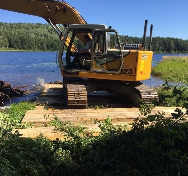 Gravemaskin jobber i vannkanten fra treunderlag, med bølger, siv og skog rundt.