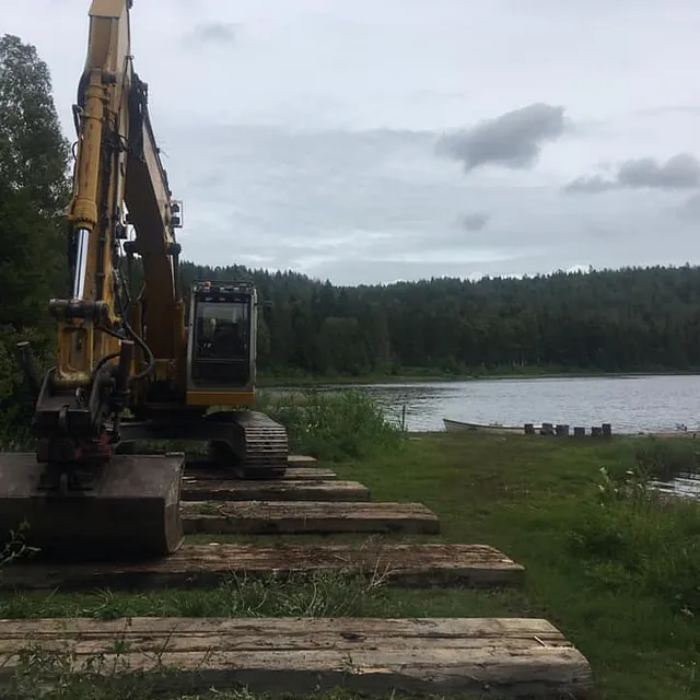 Beltegraver står på treplater ved innsjø og arbeider langs vannkanten med skog i bakgrunnen.
